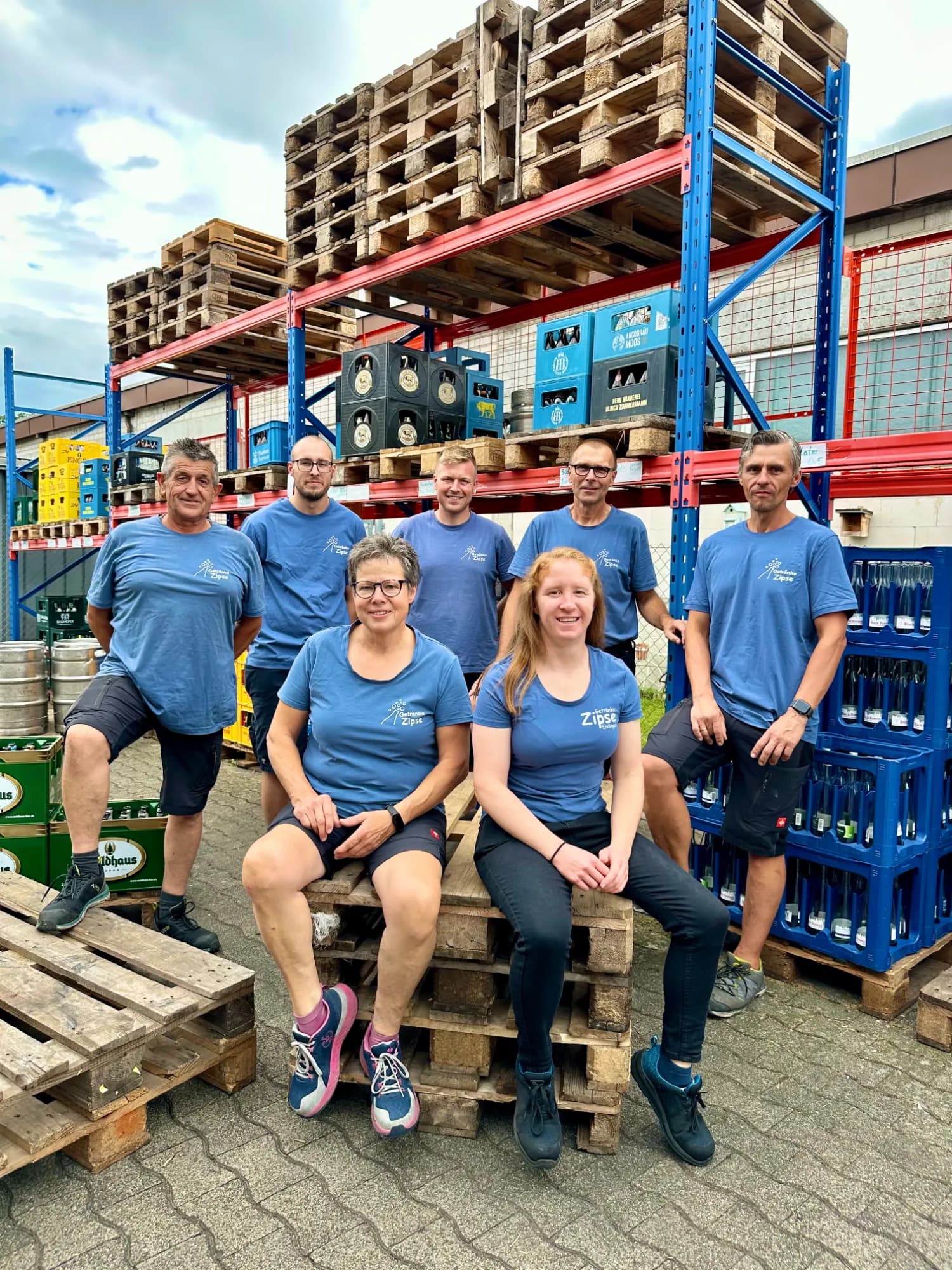 Sechs Personen in blauen T-Shirts sitzen und stehen zwischen Getränkekästen und Paletten.